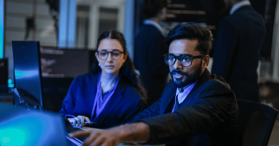 Two AI engineers collaborate in a modern tech workspace, analyzing data and discussing code on a computer screen.